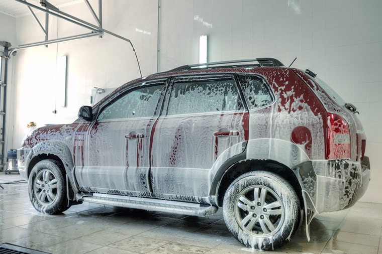 A maroon SUV covered in white soap suds is undergoing a car wash inside a brightly-lit garage. The car is parked on a tiled floor, with wall lights and a partially visible open garage door in the background.