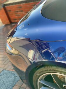 Close-up of a shiny blue car's rear end, highlighting the taillight, sleek curved design, and tire. The car's polished surface reflects nearby brick walls, blue sky, and surrounding environment. The vehicle is parked on a paved driveway.