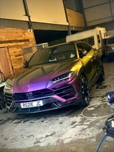 A metallic color-shifting Lamborghini Urus is parked inside a dimly lit garage. The luxury SUV features a personalized license plate reading "RO21E JC." The background includes wooden panels and other vehicles, including a white caravan.