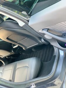 The image shows the interior of a car, focusing on the area between the front seats and the back seats. The car seats are upholstered in gray leather, and the floor is covered with black-striped mats. The door panel shows part of the armrest and side compartment.