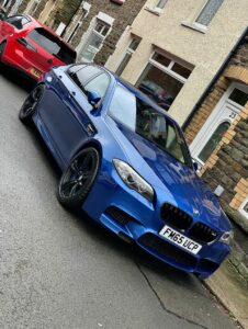 A blue BMW is parked on a street in front of a row of houses. A red car is parked behind it. The houses have stone facades and white-framed doors and windows. The license plate on the BMW reads "FM65 UCP." The street appears to be slightly wet.