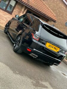 A black Range Rover with tinted windows is parked on a driveway near a brick house with a tiled roof. The vehicle’s license plate reads "J054 SHJ." The surface appears wet as if from recent rain. A satellite dish is visible on the house.