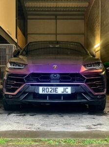 A front-facing view of a Lamborghini Urus in a garage, displaying a gradient purple to black color. The car's grille and headlights are prominent, and the license plate reads "RO21E JC". The garage background features some industrial elements and lights.