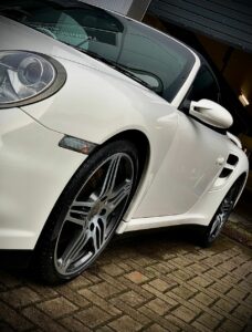 A sleek white sports car is parked on a brick pavement. The image focuses on the front left side, highlighting the shiny alloy wheel and the car's aerodynamic design. The background includes a partially open garage door.