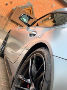 Close-up view of a sleek, matte gray sports car. The focus is on the rear wheel, showcasing the black rim and Pirelli tire. The background includes a red brick building with a wooden canopy and reflective windows.