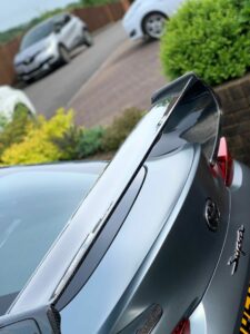 Close-up of the rear of a gray sporty car featuring a large carbon fiber rear spoiler. The background shows a paved driveway with a few other cars and green bushes near a brick wall. The scene appears to be in a residential area.