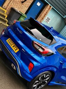 A blue Ford car is parked near a building with a blue door. The car's license plate reads "EG22 RTX." There are a green garbage bin and a yellow set of stairs nearby. A sign on the wall by the door reads, "Medical Record Storage Holders Only.