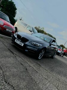 A gray BMW, parked on a paved surface, is prominent in the foreground with the license plate "RF17 ZKD." Behind it is a red Mini Cooper, a white SUV, and several other cars. Overcast sky and a mix of trees and buildings are visible in the background.