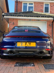 A dark blue Porsche sports car is parked in front of a brick house with a white garage door. The car has a yellow license plate that reads "2 FJY." The house's eaves are decorated with string lights. The driveway consists of light brown bricks.