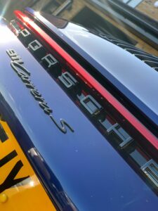 Close-up of the rear end of a blue Porsche 911 Carrera S, featuring the Porsche logo and red taillight strip above the license plate. The image is angled, showing the reflective surface and part of the vehicle's surroundings.