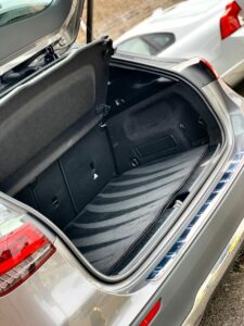 Open trunk of a silver car. The trunk area is empty and clean, with a black interior. The car is parked on the side of a street, with another white vehicle nearby. The trunk lid is open, revealing the spacious cargo area.