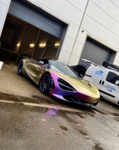 A metallic chameleon-colored sports car with a gradient of purples, blues, and golds is parked in front of a white industrial building with large roller doors. A white van with business logos is parked beside the car. The ground appears to be wet.