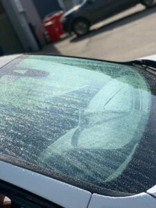 Close-up of a car's windshield covered in water droplets, reflecting sunlight. A red trash bin and another parked car are blurred in the background. The windshield wipers are visible beneath the water-covered glass.