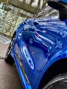 Close-up of a shiny blue car with reflections of trees and another vehicle on its polished surface. The photo is taken at an angle from the rear side, showing the car parked on a wet road near a stone wall and a metal fence.