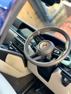 Close-up of a car's interior featuring a steering wheel with the Porsche logo, dashboard controls, a touchscreen display, and beige leather seats. The steering wheel has various buttons, while the dashboard displays modern, sleek design elements.