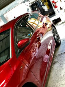 A shiny red car is parked indoors beside a white van with its back doors open. The reflection of the surrounding environment is clearly visible on the car's surface. The car appears to be clean and polished.