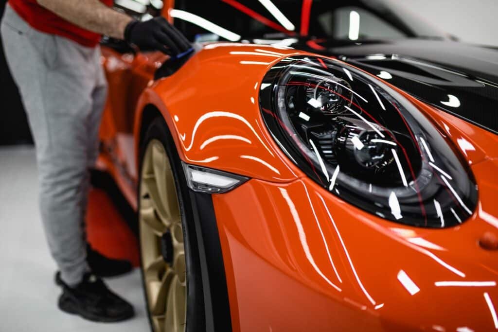 Close-up of a bright orange sports car being detailed by a person wearing a red shirt and gray pants, with black gloves. The car has sleek lines, prominent headlights, and gold-colored rims. The reflective surface shows lighting and the surrounding environment.