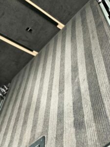 A close-up view of a carpeted floor with alternating light and dark gray stripes. The patterned carpet creates a subtle linear design. The floor is indoors, and part of a wall with a square cutout can be seen in the background.