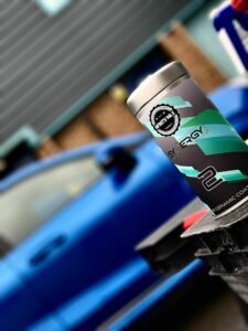 A close-up view of a bottle with a camouflaged green and black design labeled "Synergy 2" resting on a black surface. In the blurred background, there is a blue car and the exterior of a building with gray siding.