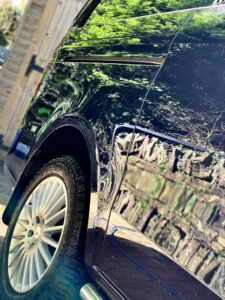 Close-up of a shiny black car parked in a driveway. The car's left side, including the rear tire, is visible, reflecting green foliage and a stone wall. The background shows a partially opened wooden gate and a lush garden. The tire's intricate design is prominent.