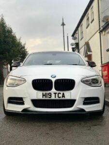 Front view of a white BMW car with license plate "H19 TCA" parked on a wet street between residential buildings. Overcast sky and tree visible in the background.