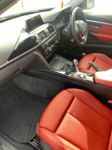 The interior of a modern car with red leather seats, a dashboard featuring a central display screen, various control buttons, a gear shift lever, and black floor mats. Sunlight illuminates the area, highlighting the sleek design and clean surfaces.