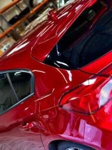 Close-up of a vibrant red car featuring the sleek rear section, including the taillight, rear window, and left-side door. The background shows shelves and various items in a garage or workshop setting.