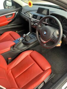 Interior of a BMW car featuring red leather seats, a black dashboard and steering wheel, a central console with gear shift, and a display screen. The car's cabin is clean and modern, with a hanging air freshener visible from the rearview mirror.