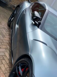 Close-up angle of a sleek, metallic silver sports car parked on a brick driveway. The image focuses on the car's side, emphasizing its aerodynamic curves, shiny exterior, and red brake calipers visible through the black alloy wheels.