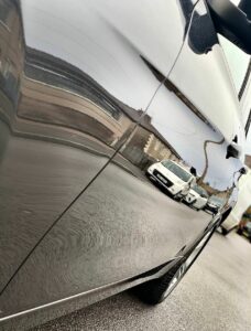 A close-up shot of a car door with a highly reflective, smooth surface. The reflection on the car door shows several buildings and a white car parked on the street. The focus is on the shiny exterior of the car, highlighting its polished and clean appearance.