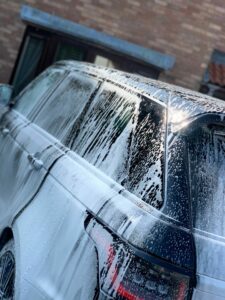 A large black SUV covered in soap suds during a car wash, with the background showing a building with brick walls and windows. The sunlight reflects off the wet surface of the SUV, highlighting the cleaning process.