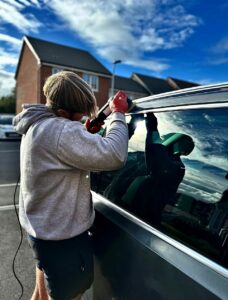 A person wearing a grey hoodie and gloves uses a tool to work on the window of a car parked outdoors. The sky is partly cloudy, and there are houses in the background. The sunlight casts a reflection on the car’s window.