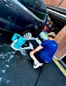 A person wearing a blue shirt and shorts is washing a dark-colored car outside a garage. They are using a hose and soapy water, with a blue bucket nearby. The garage door is open, revealing a cluttered interior with various items.