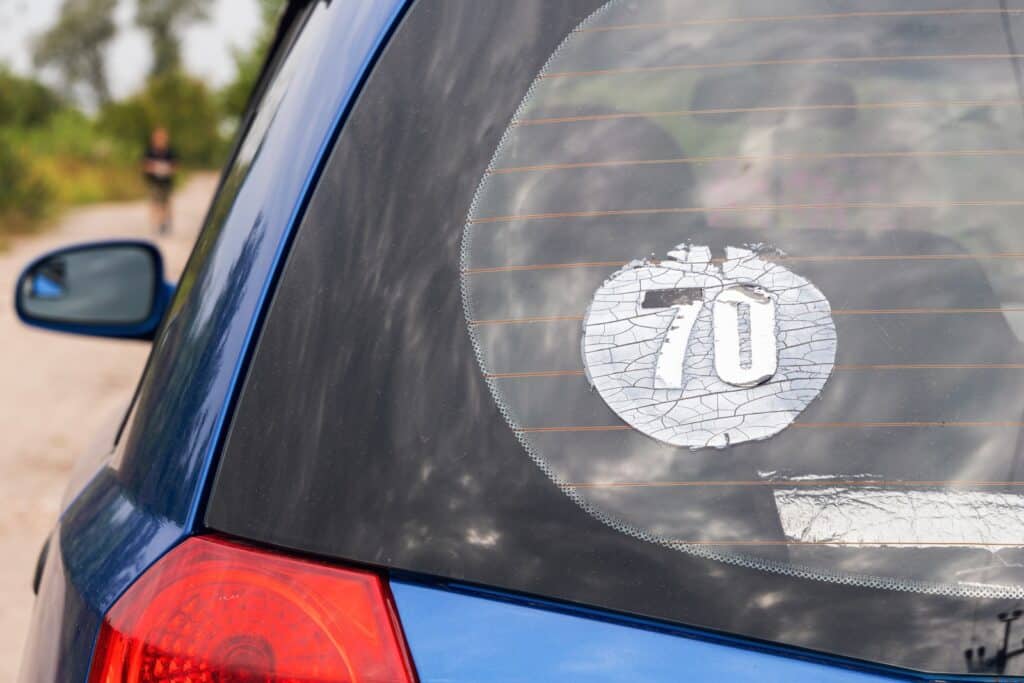 A close-up of the rear window on a blue car features a large, cracked sticker displaying the number 70. Meanwhile, in the background, amidst lush greenery, a blurred figure walks along a dirt road. It seems like this sticker is due for removal to restore the car's sleek look.