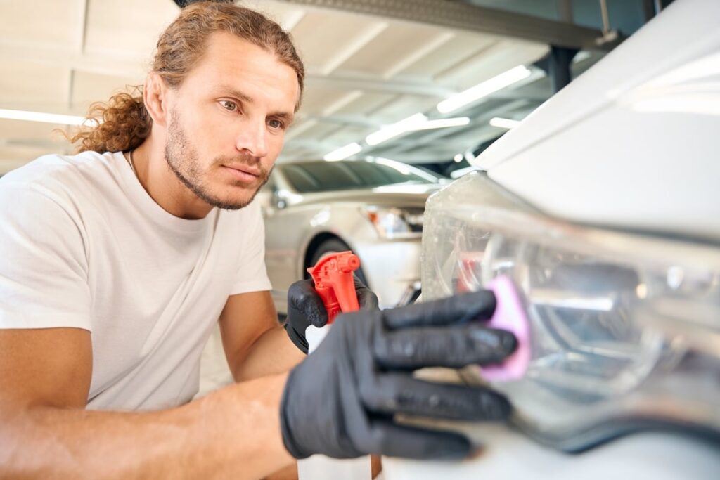 A person with long hair and a white shirt, wearing black gloves, is cleaning a car's headlight using a cloth. They are focused on the task in a well-lit garage, with other cars visible in the background.