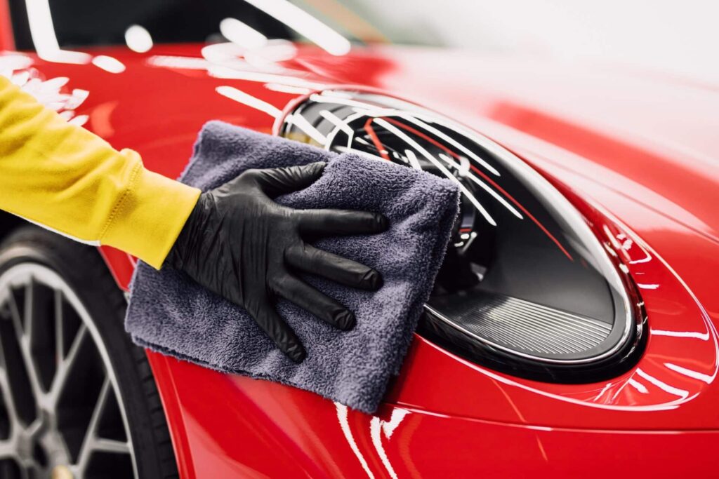 A person wearing a yellow sleeve and black glove is polishing the headlight of a red sports car with a gray microfiber cloth. The car is shiny and well-maintained.