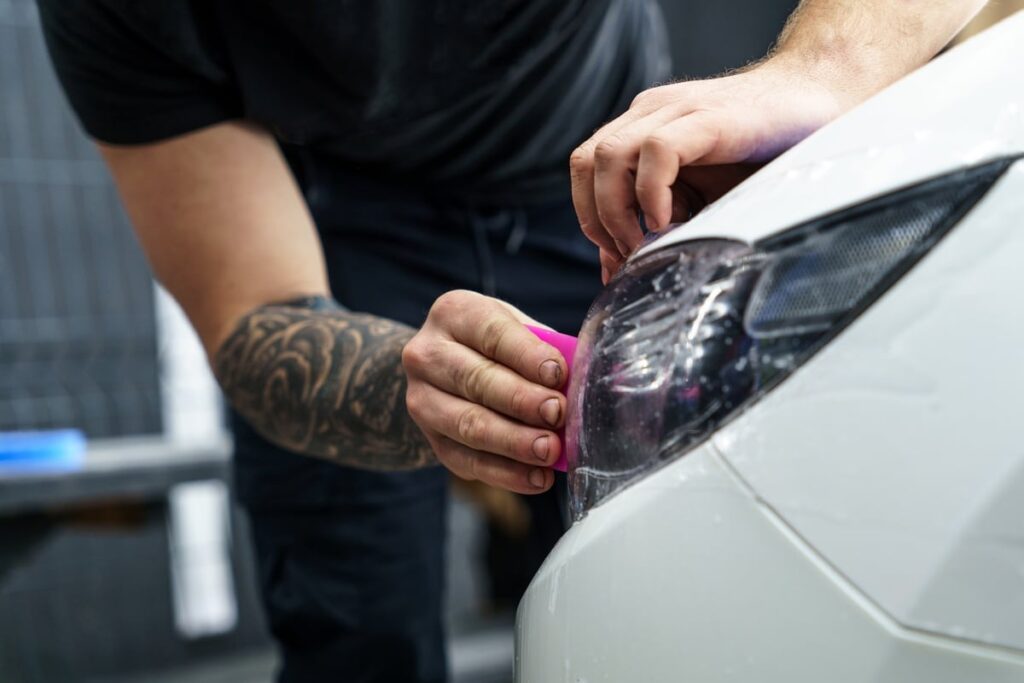 A person with a tattooed arm applies a protective film to the headlight of a white car, using a pink tool. They are focused on ensuring a smooth application, highlighting the detailing work.